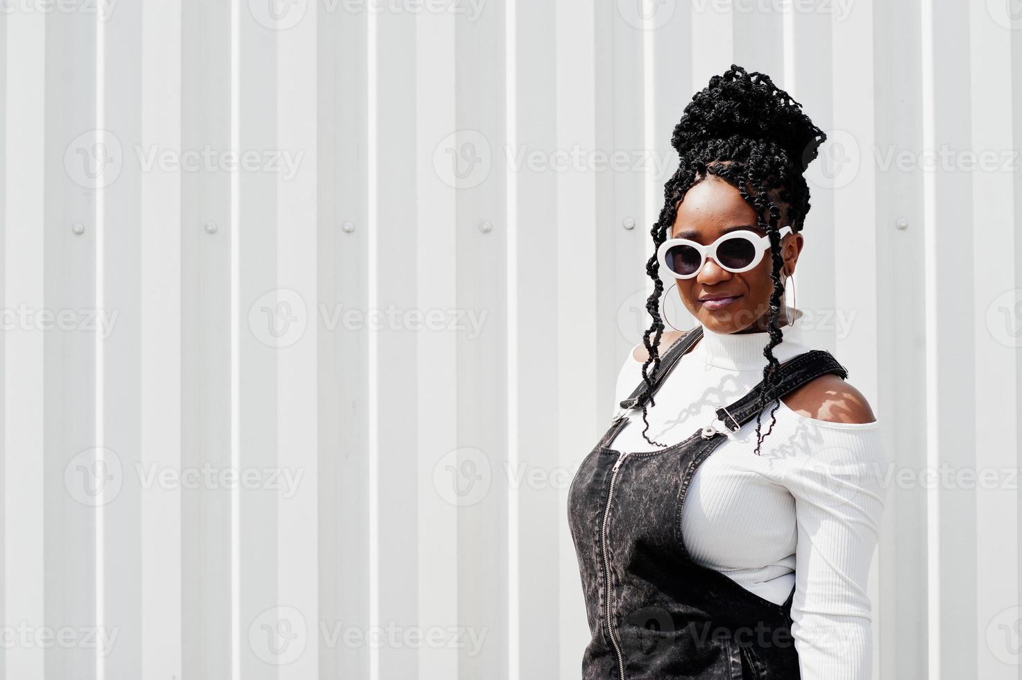 mulher africana em saia de macacão jeans, óculos de sol brancos posou contra a parede de aço branco. foto