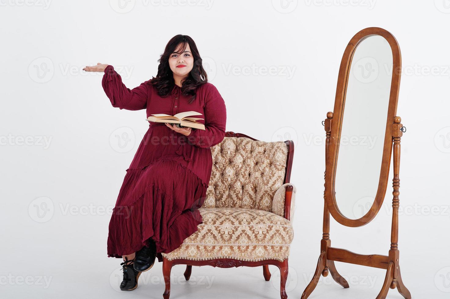 mulher atraente do sul da Ásia em vestido vermelho profundo posou no estúdio em fundo branco contra espelho e cadeira, leia um livro. foto