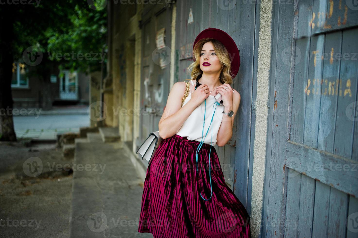 menina modelo loira elegante e bonita na saia de veludo vermelho elegante, blusa branca e chapéu, posou com telefone e fones de ouvido. foto