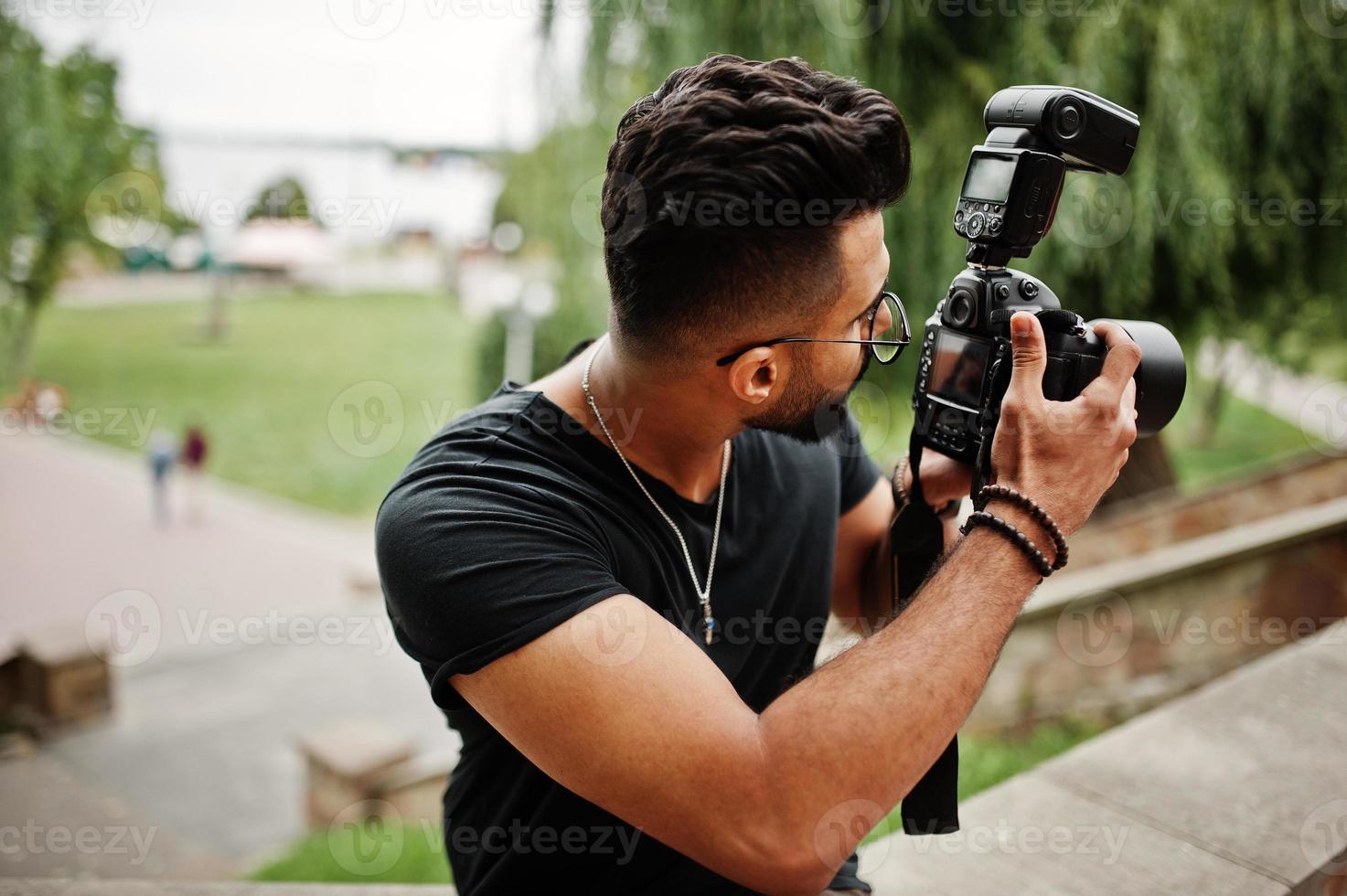 Fotógrafo de homem macho de barba árabe linda e bonita em óculos e camiseta preta com câmera profissional nas mãos. foto