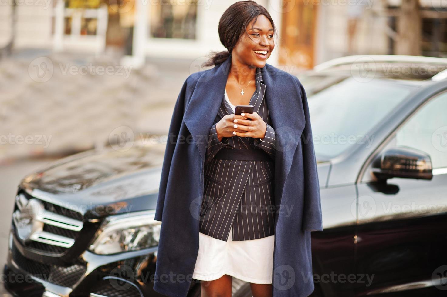 sucesso elegante mulher afro-americana de casaco contra carro suv de negócios preto com telefone celular nas mãos. foto