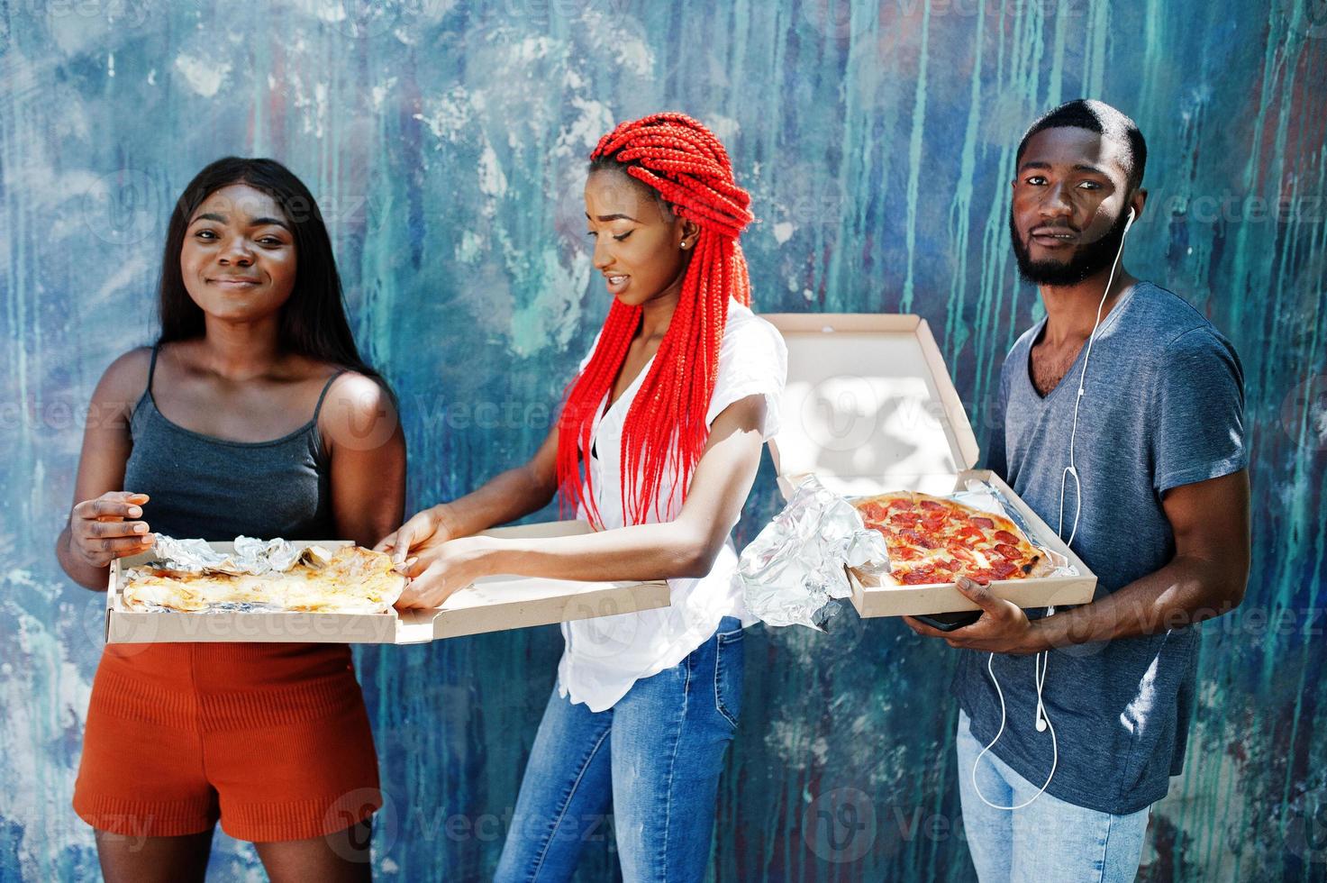 três alegres amigos afro-americanos segurando caixas de pizza contra a parede de pintura. foto