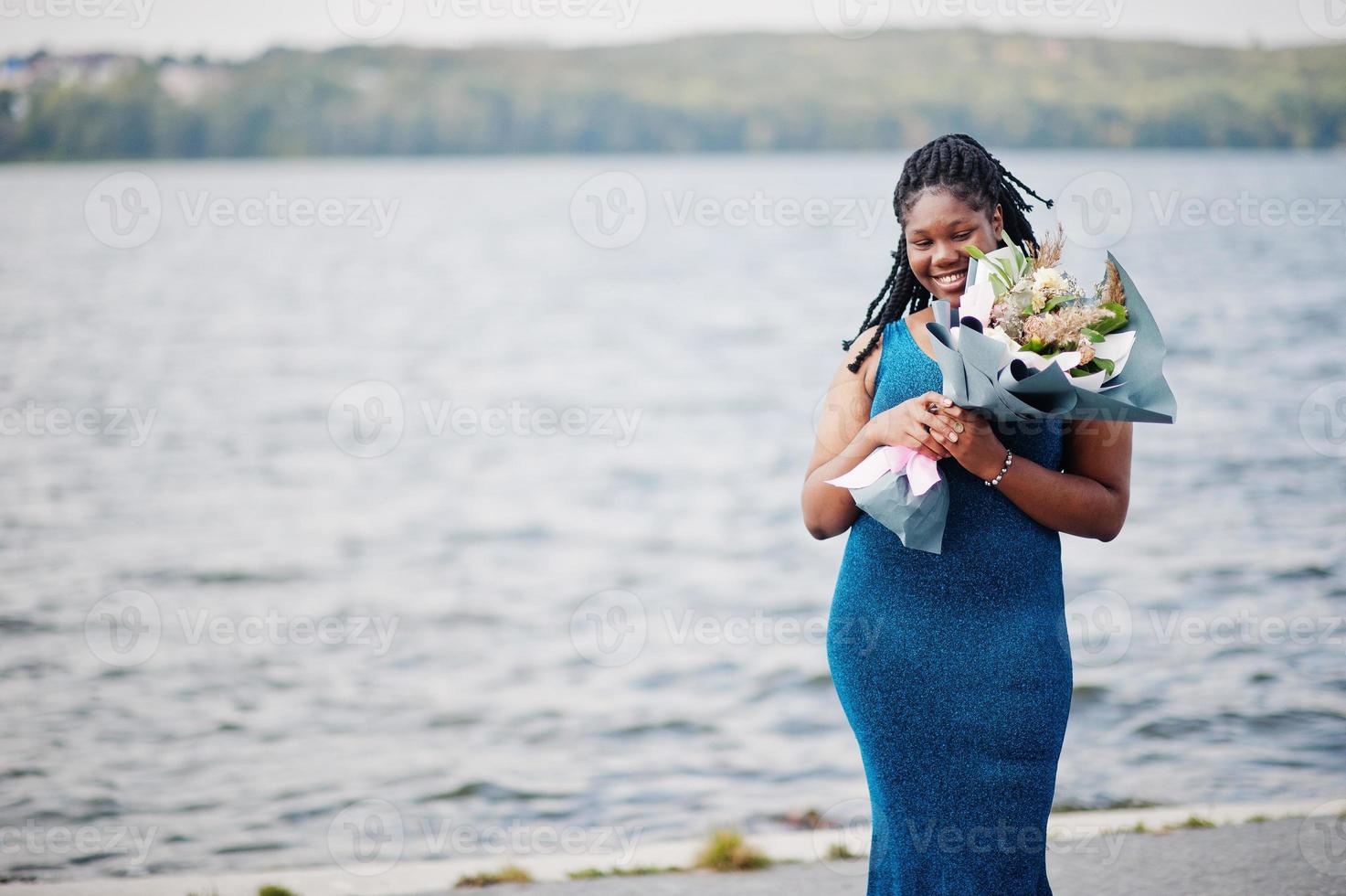 modelo plus size de pele escura afro-americana posou em um vestido azul brilhante com buquê de flores contra o lado do mar. foto