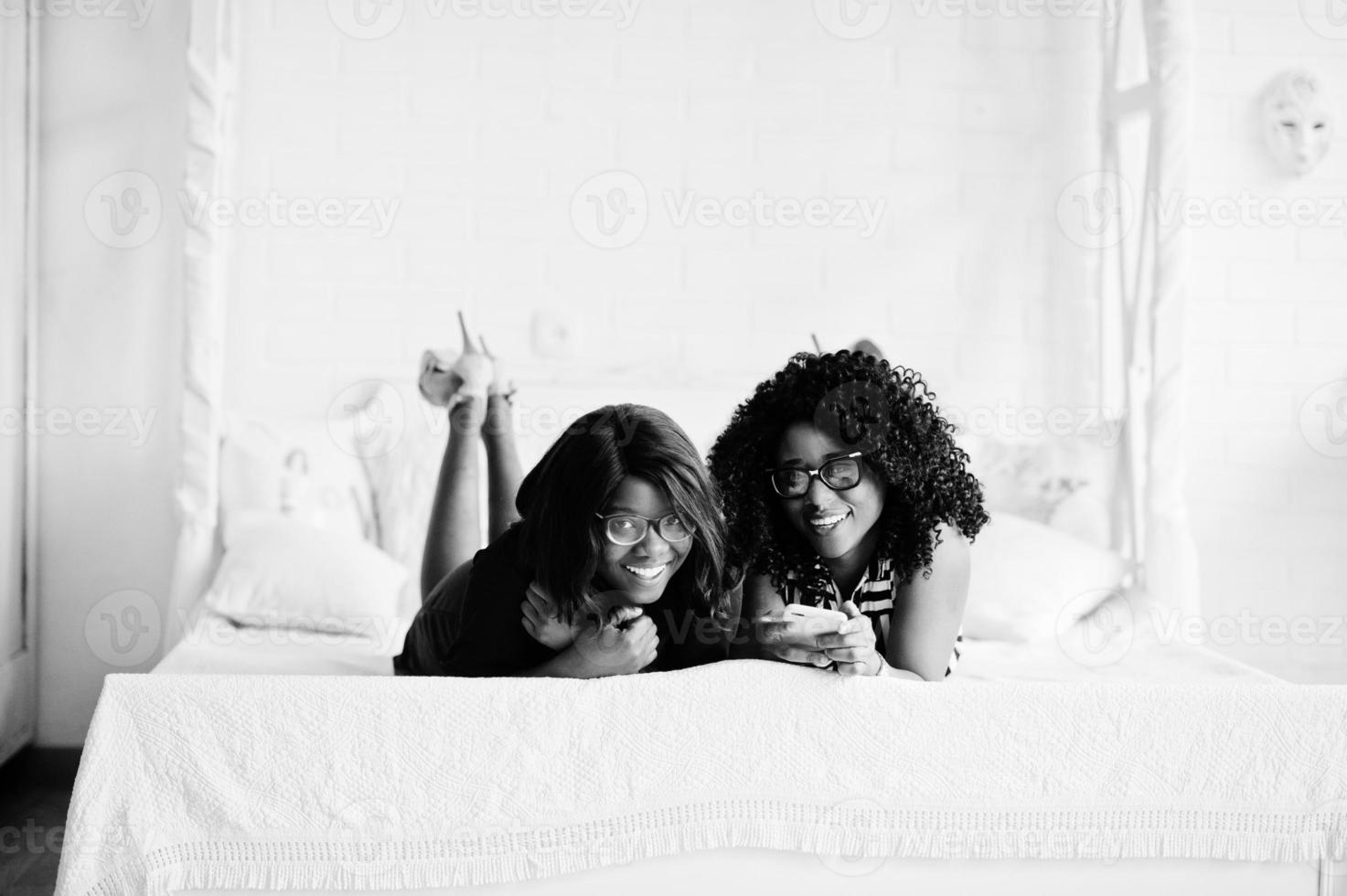 duas amigas africanas usam óculos deitadas na cama do quarto branco interno e olhando para o celular. foto