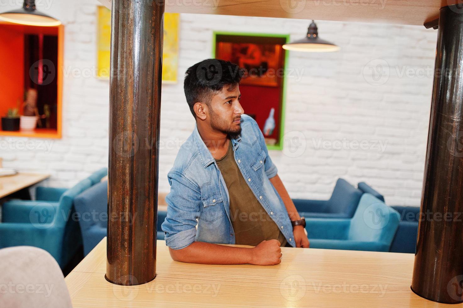 retrato de bonito bem sucedido barbudo sul-asiático, jovem freelancer indiano em pé de camisa jeans azul no café. foto