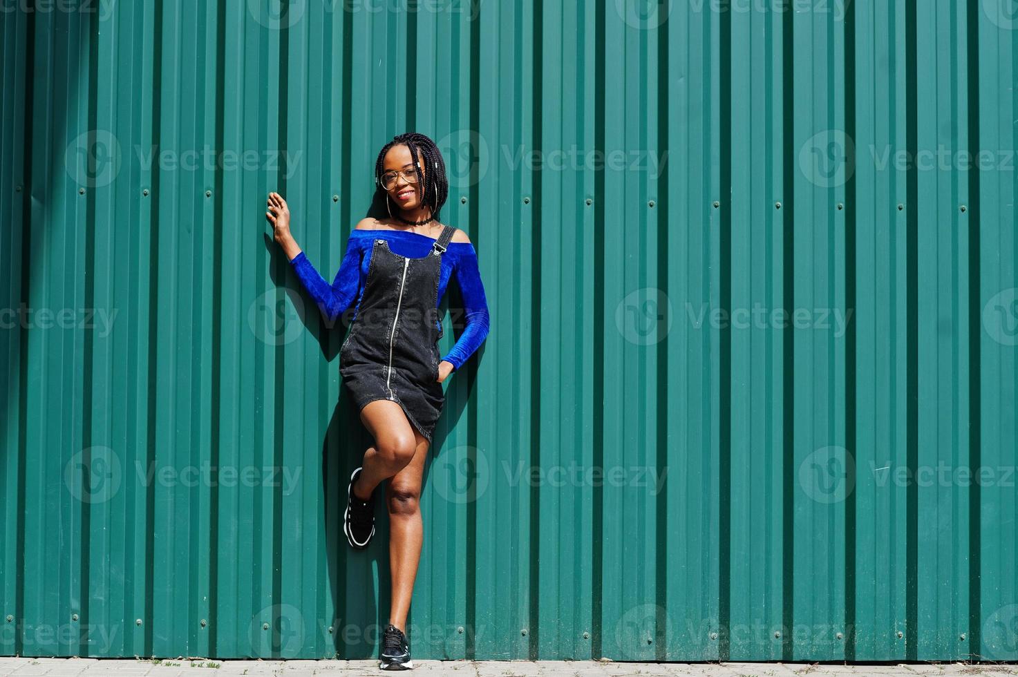 mulher africana em saia de macacão jeans, óculos posou contra a parede de aço verde. foto