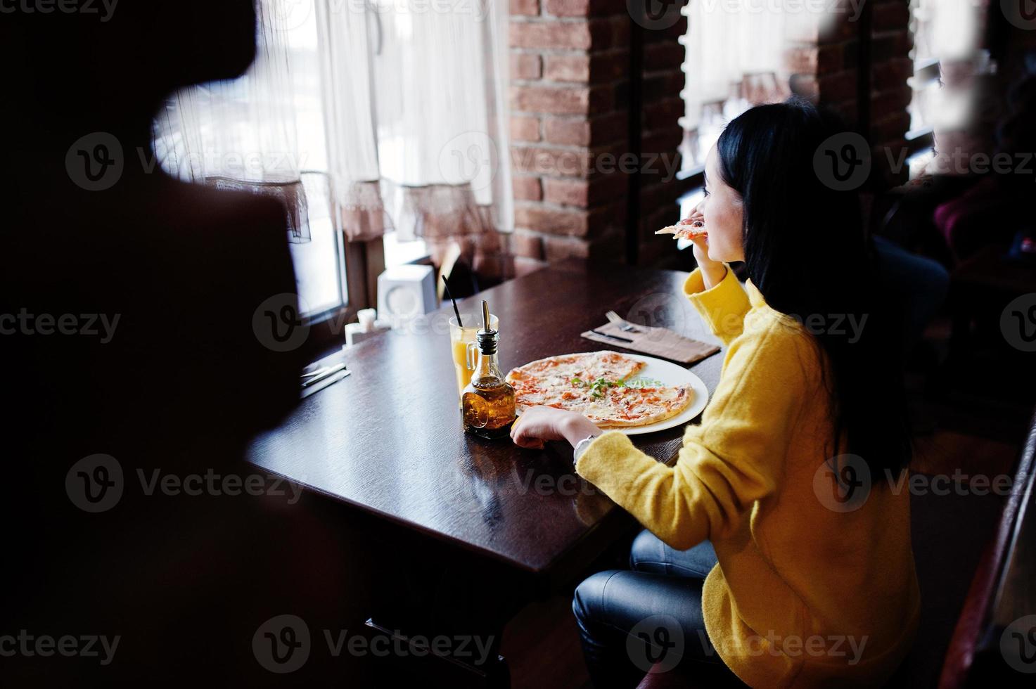 menina morena engraçada de suéter amarelo comendo pizza no restaurante. foto