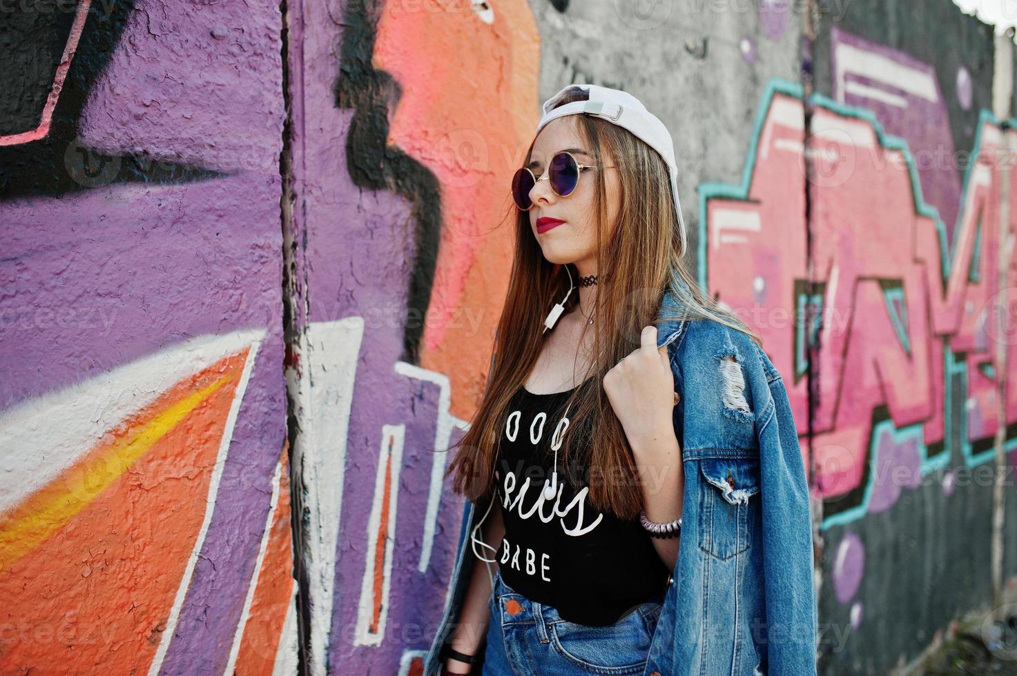 menina elegante casual hipster no boné, óculos escuros e calça jeans, ouvindo música de fones de ouvido do telefone celular contra grande parede de graffiti. foto