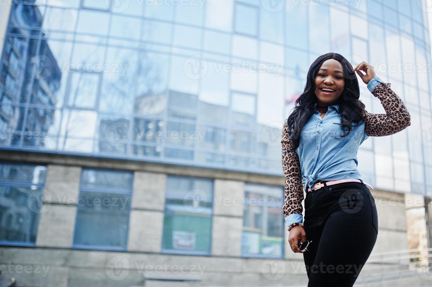 garota afro-americana hipster vestindo camisa jeans com mangas de leopardo posando na rua contra o prédio de escritórios moderno com janelas azuis. foto