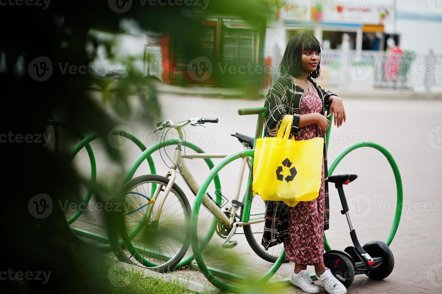linda mulher afro-americana em pé perto de segway ou hoverboard e bicicleta. garota negra com símbolo de reciclagem de sacos ecológicos de pano amarelo. foto