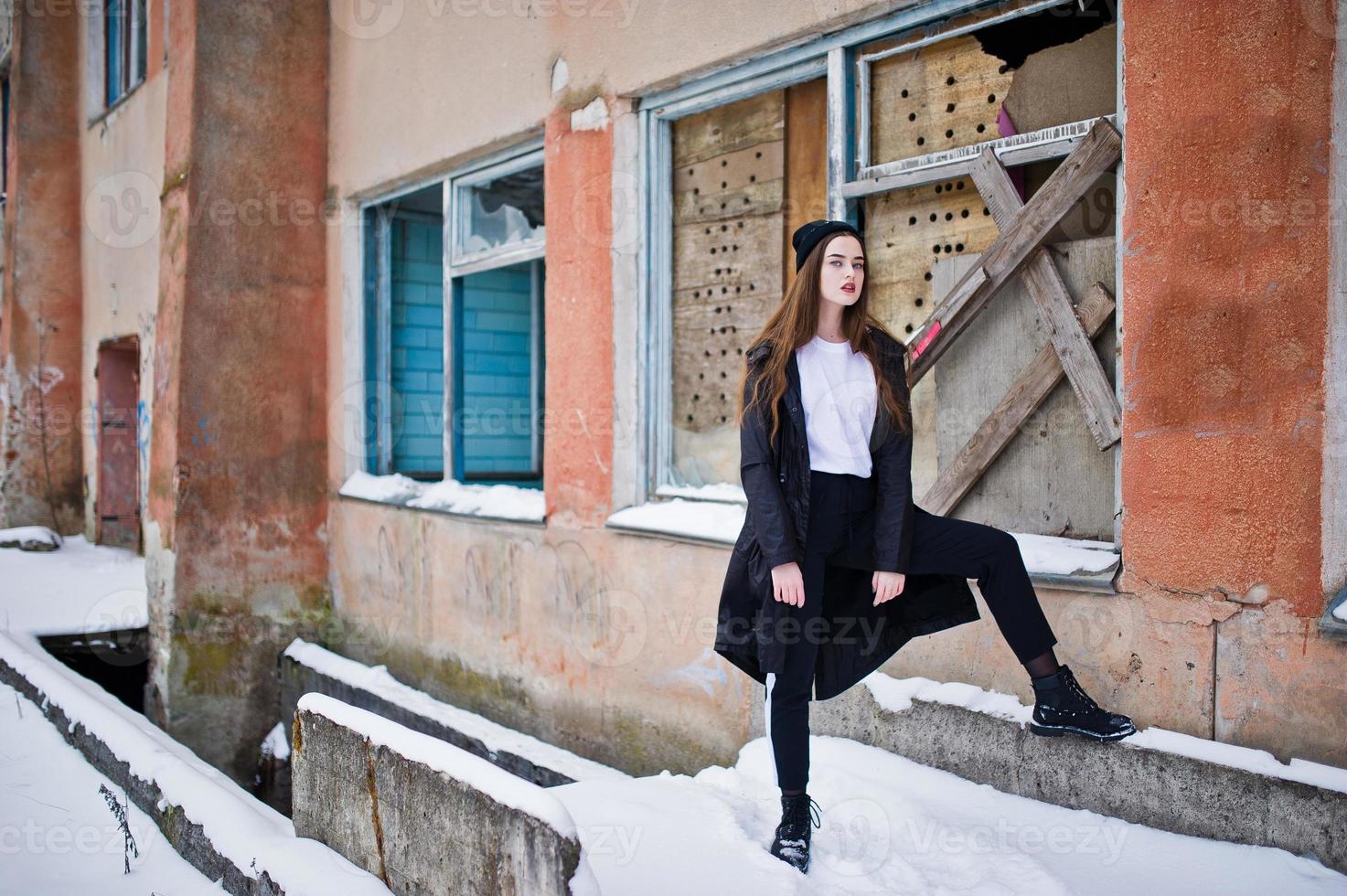 modelo morena de pernas longas na moda no manto preto longo posou ao ar livre no dia de inverno contra a velha parede do grunge com janelas quebradas. foto