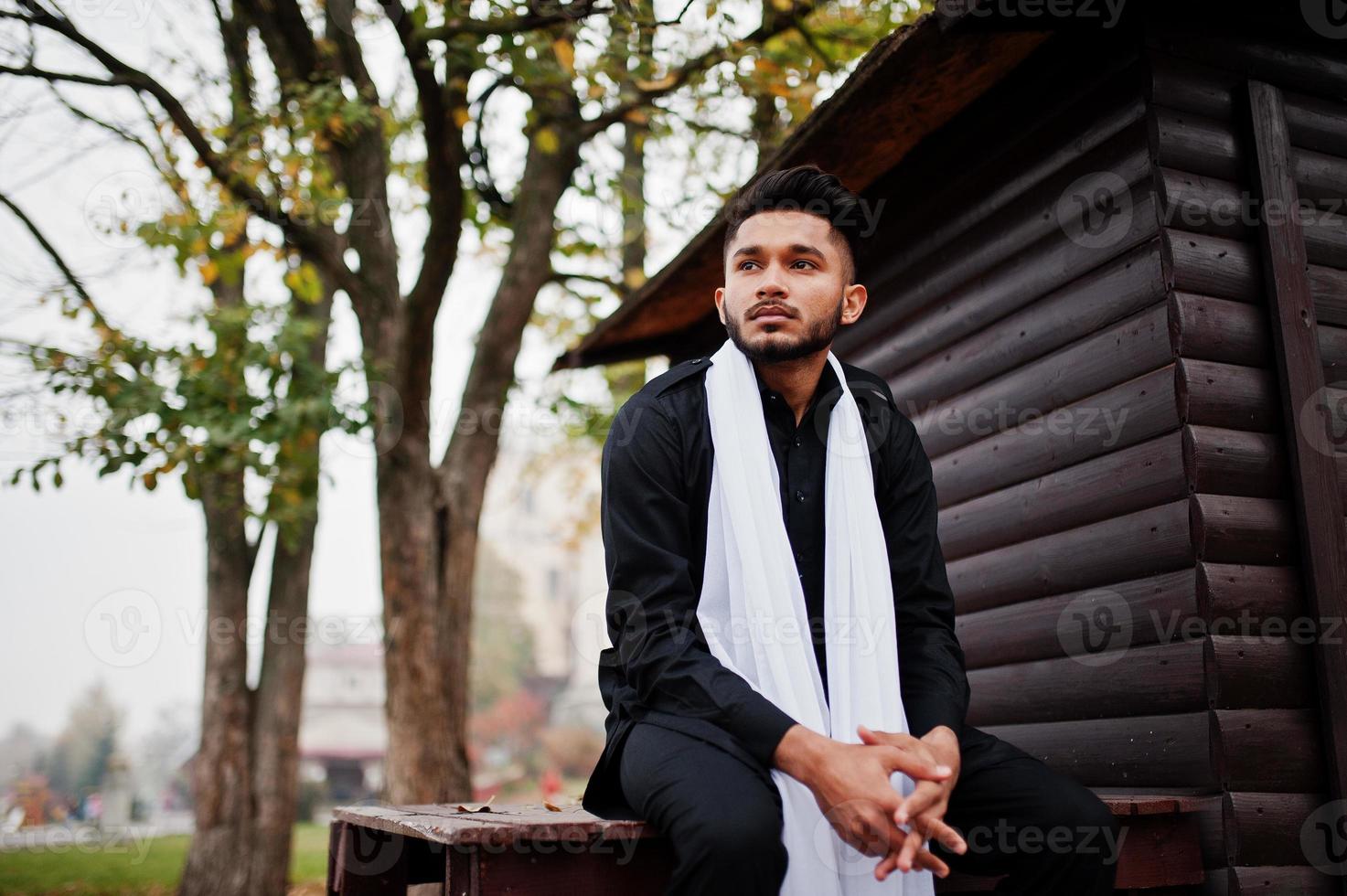 homem elegante indiano em roupas tradicionais pretas com lenço branco posou ao ar livre contra um fundo de madeira. foto