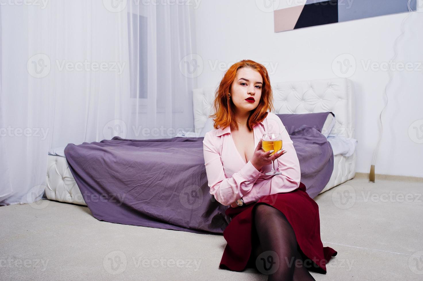 linda garota ruiva de blusa rosa e saia vermelha com copo de vinho na mão perto da cama no quarto. foto