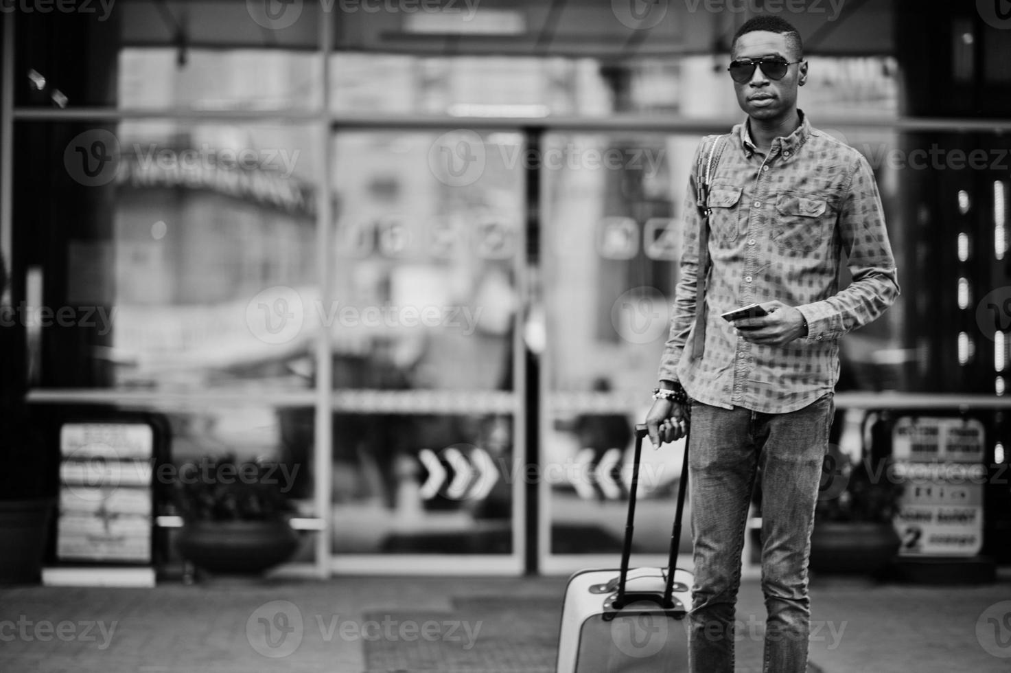 homem afro-americano de camisa quadriculada, com mala e mochila. viajante negro contra a rodoviária. foto