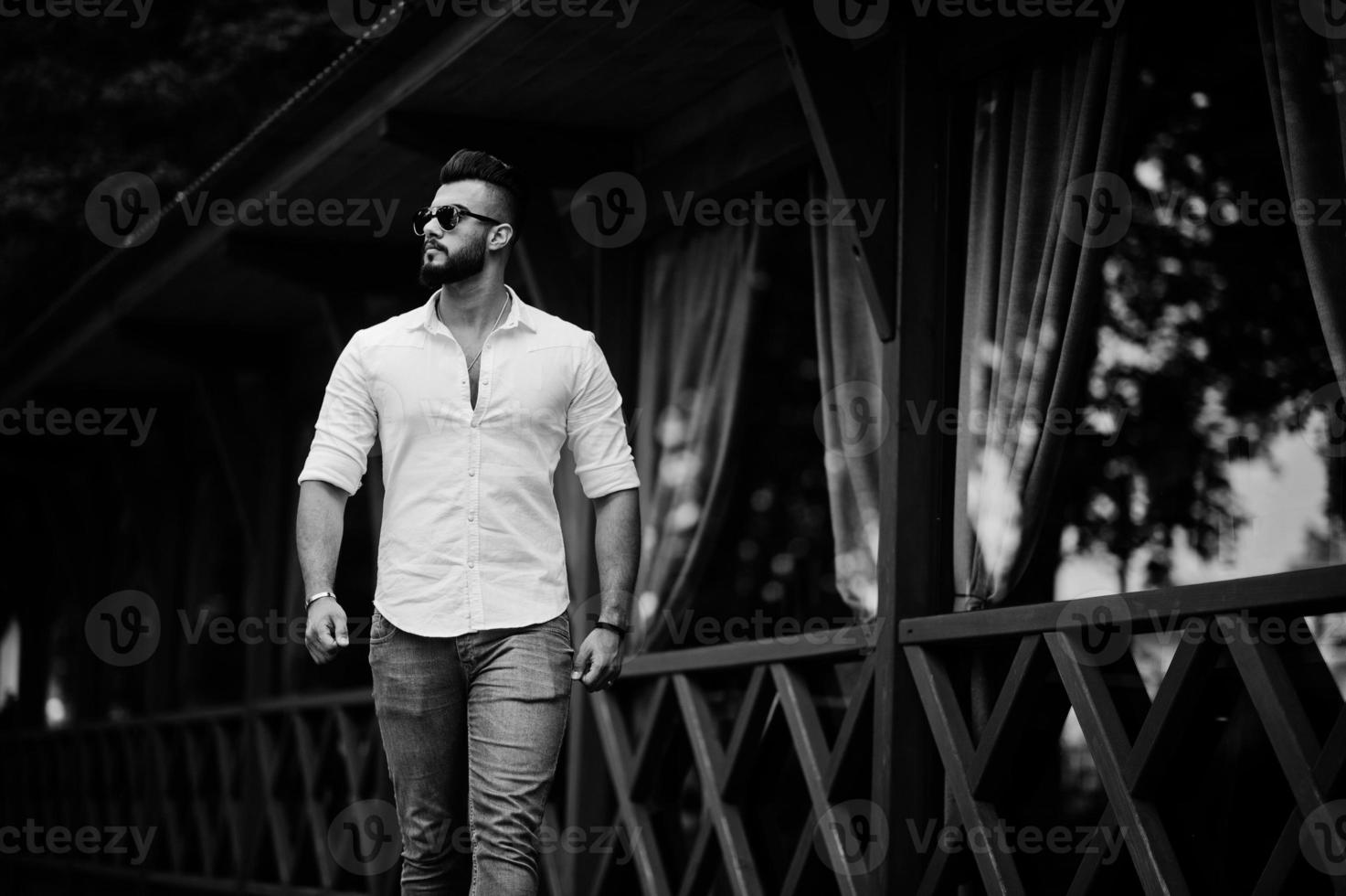 elegante modelo de homem árabe alto na camisa branca, jeans e óculos de sol andando na rua da cidade. barba cara árabe atraente. foto