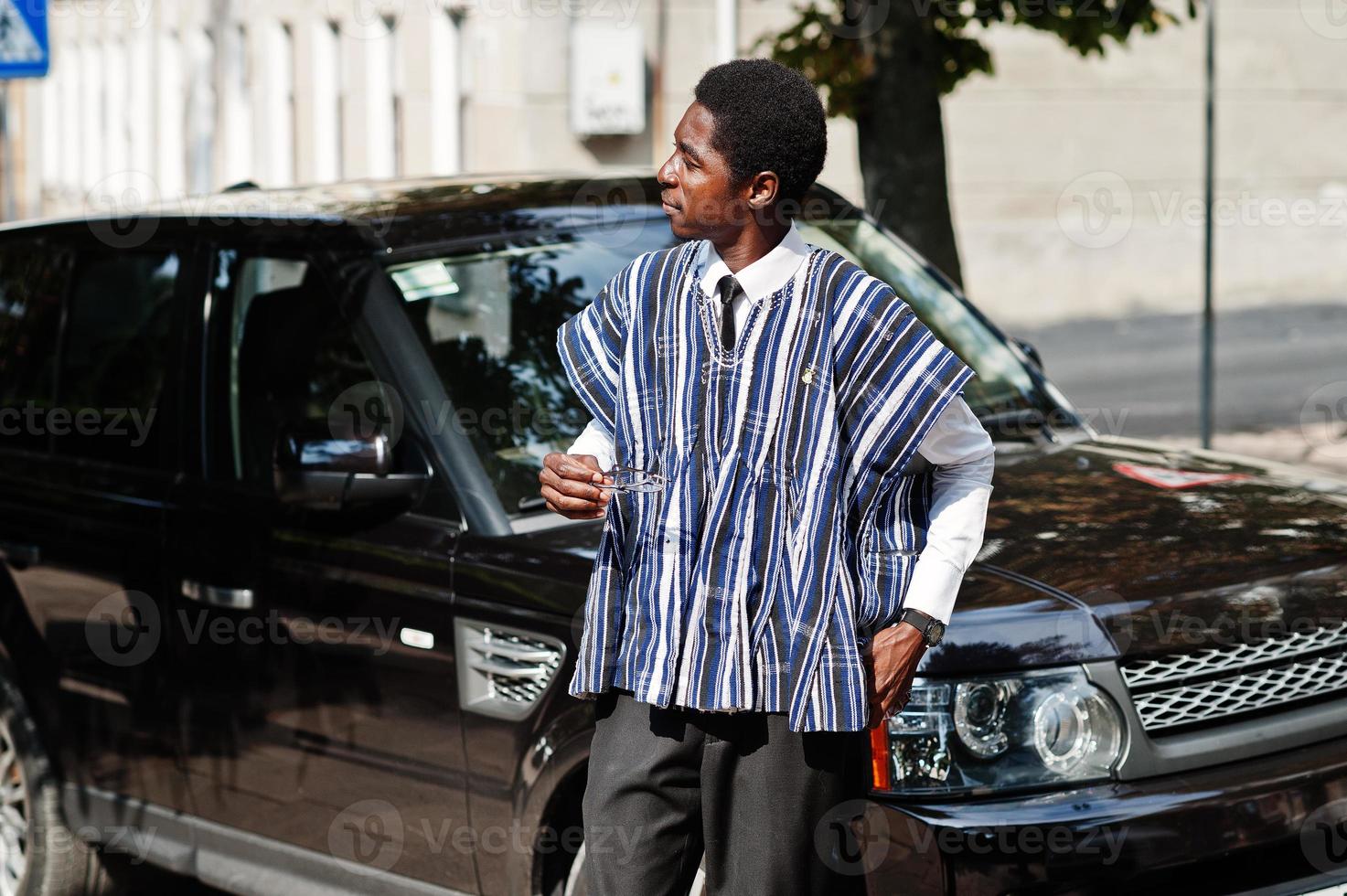 empresário africano em roupas tradicionais e óculos com celular contra carro preto suv. ricos africanos. foto