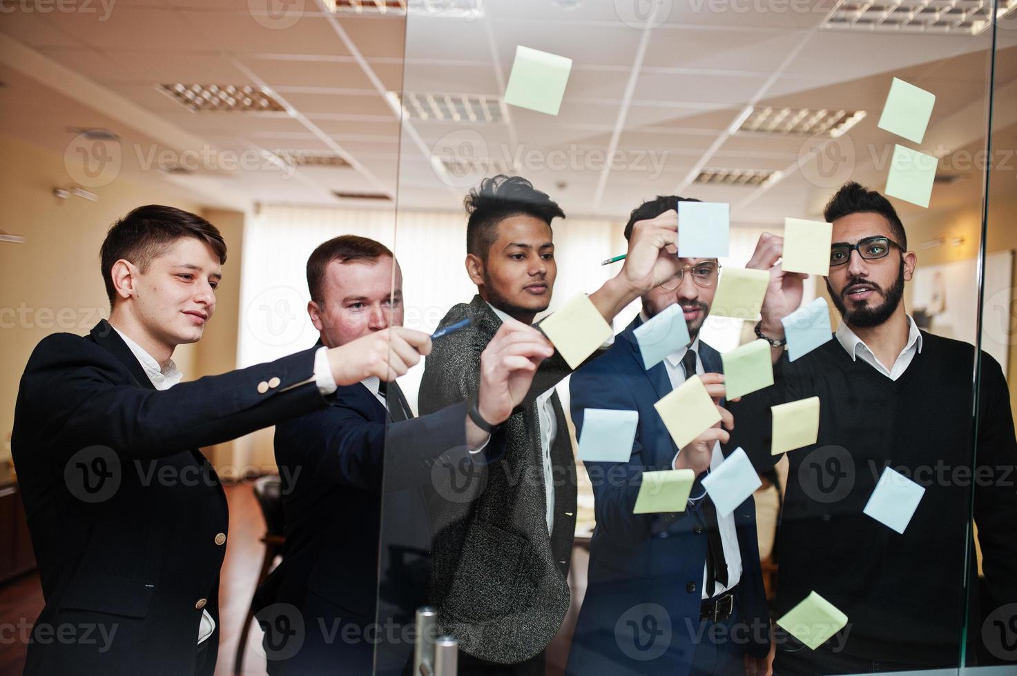 homem de negócios apontando no vidro com notas de papel colorido. grupo diversificado de funcionários do sexo masculino em trajes formais usando adesivos. foto