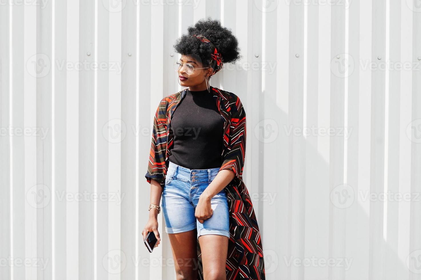 mulher africana com cabelo afro, em shorts jeans e óculos posou contra a parede de aço branca com o celular na mão. foto
