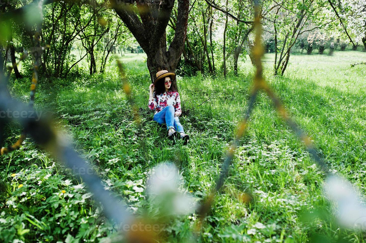 retrato de primavera de menina morena de óculos cor de rosa e chapéu no jardim de flor verde. foto