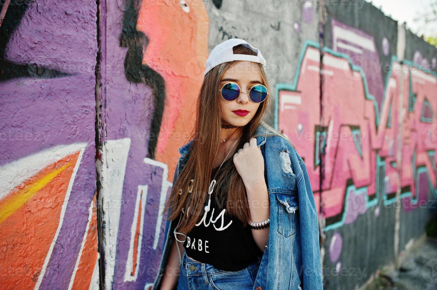 menina elegante casual hipster no boné, óculos escuros e calça jeans, ouvindo música de fones de ouvido do telefone celular contra grande parede de graffiti. foto