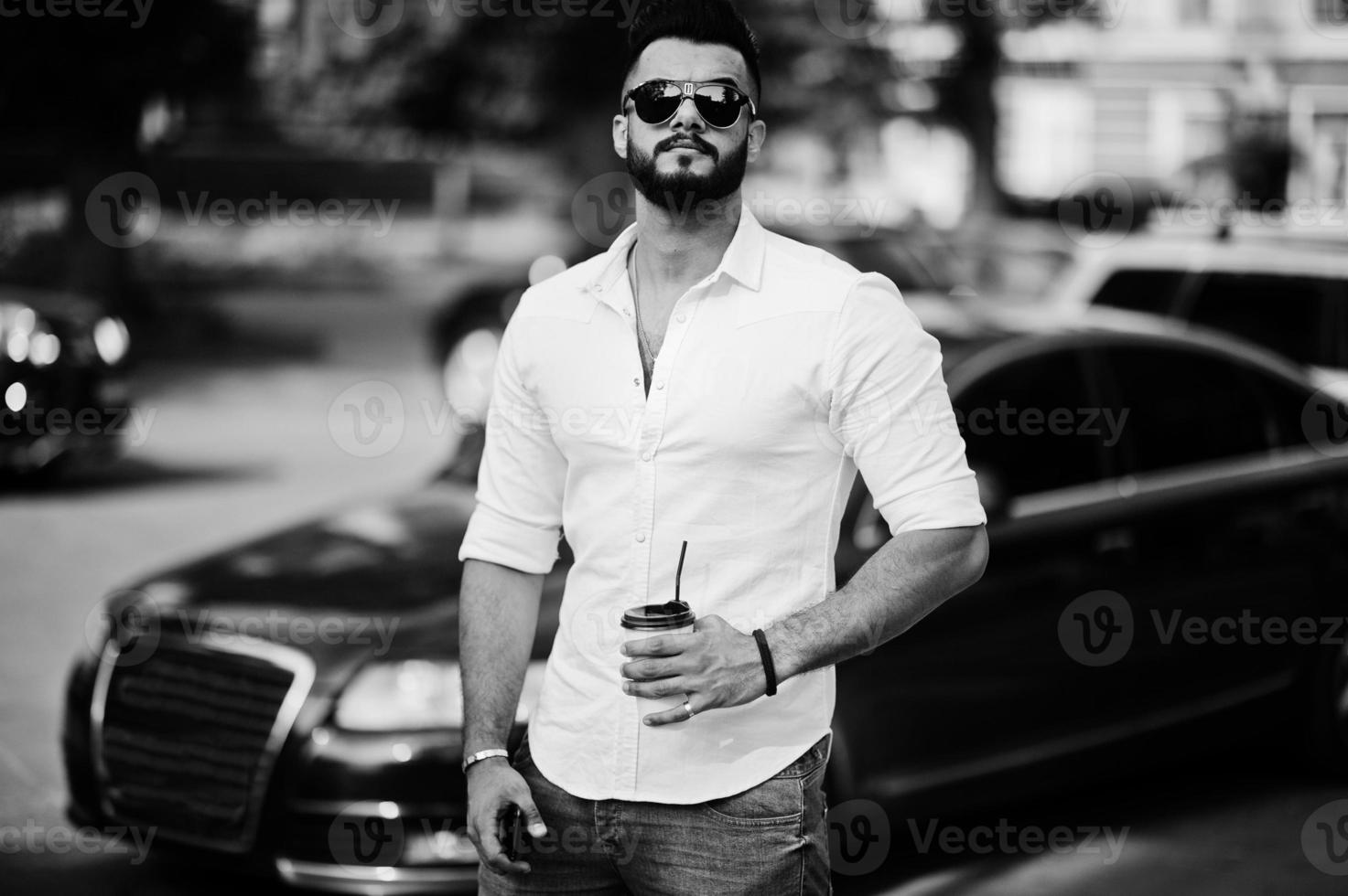 elegante modelo de homem árabe alto na camisa branca, jeans e óculos de sol posou na rua da cidade. barba cara árabe atraente com café contra carro de negócios preto. foto