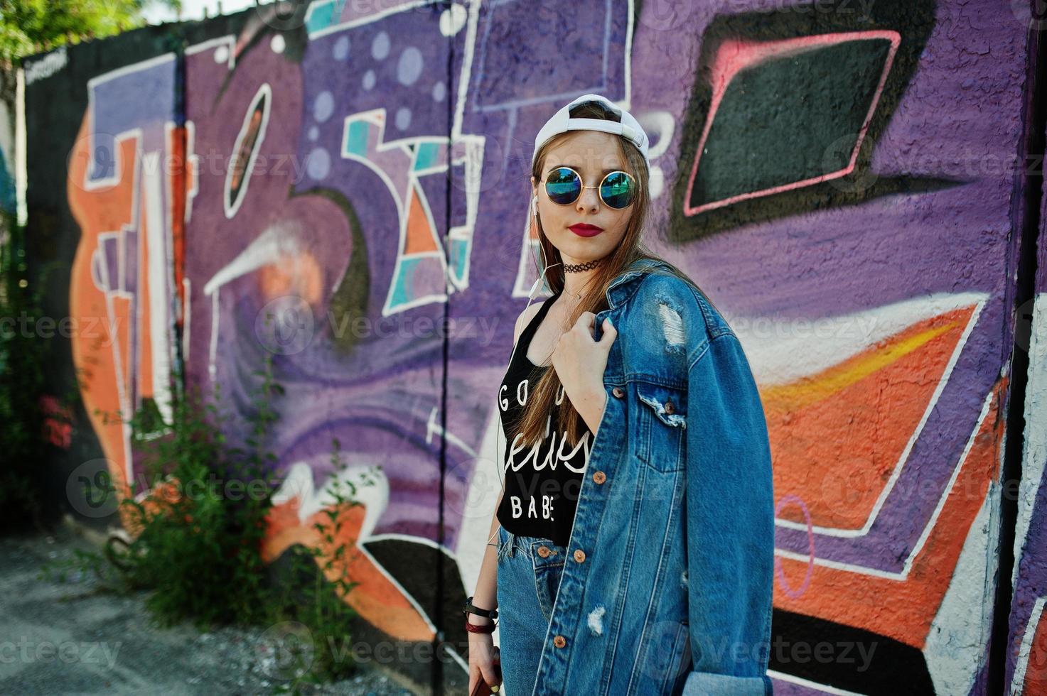 menina elegante casual hipster no boné, óculos escuros e calça jeans, ouvindo música de fones de ouvido do telefone celular contra grande parede de graffiti. foto