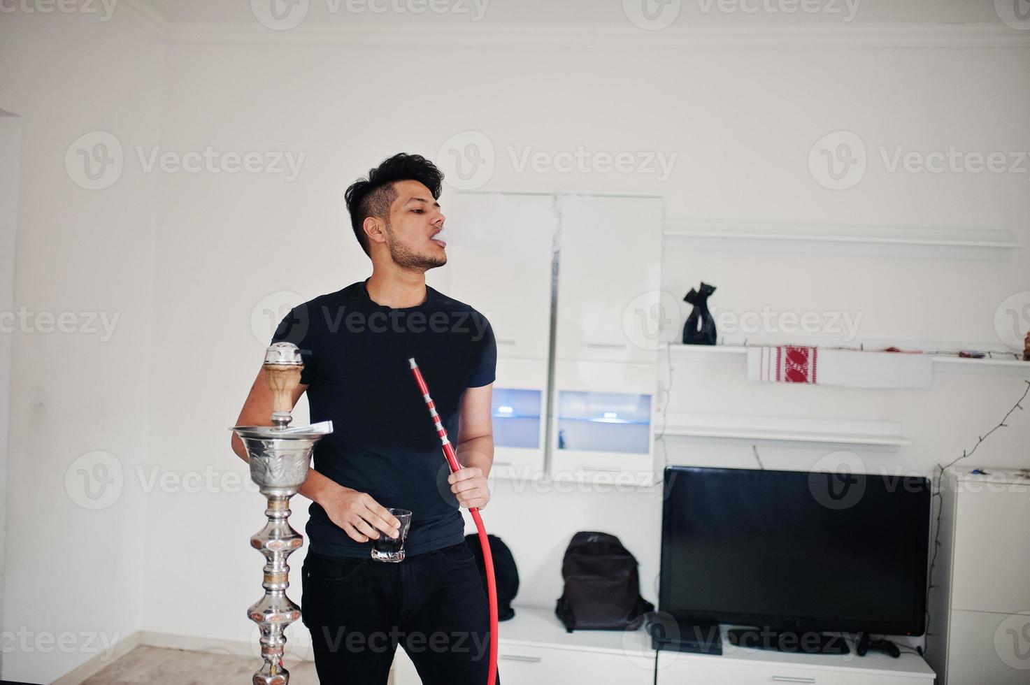 homem indiano bonito e elegante em pé de preto no quarto, fumando cachimbo de água com óculos na mão. foto