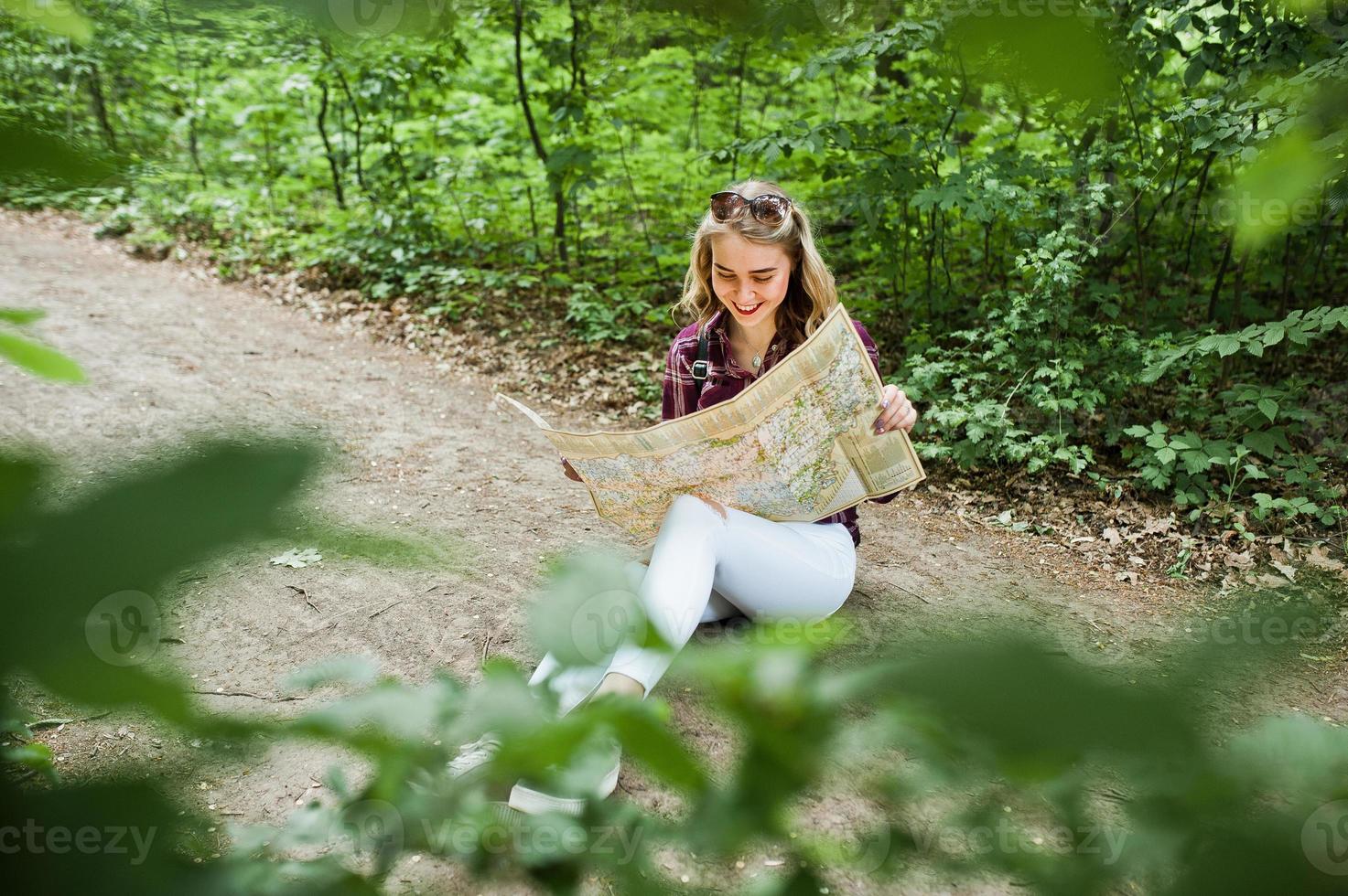 retrato de uma loira linda jovem positiva sentada no chão com um mapa nas mãos na floresta. foto