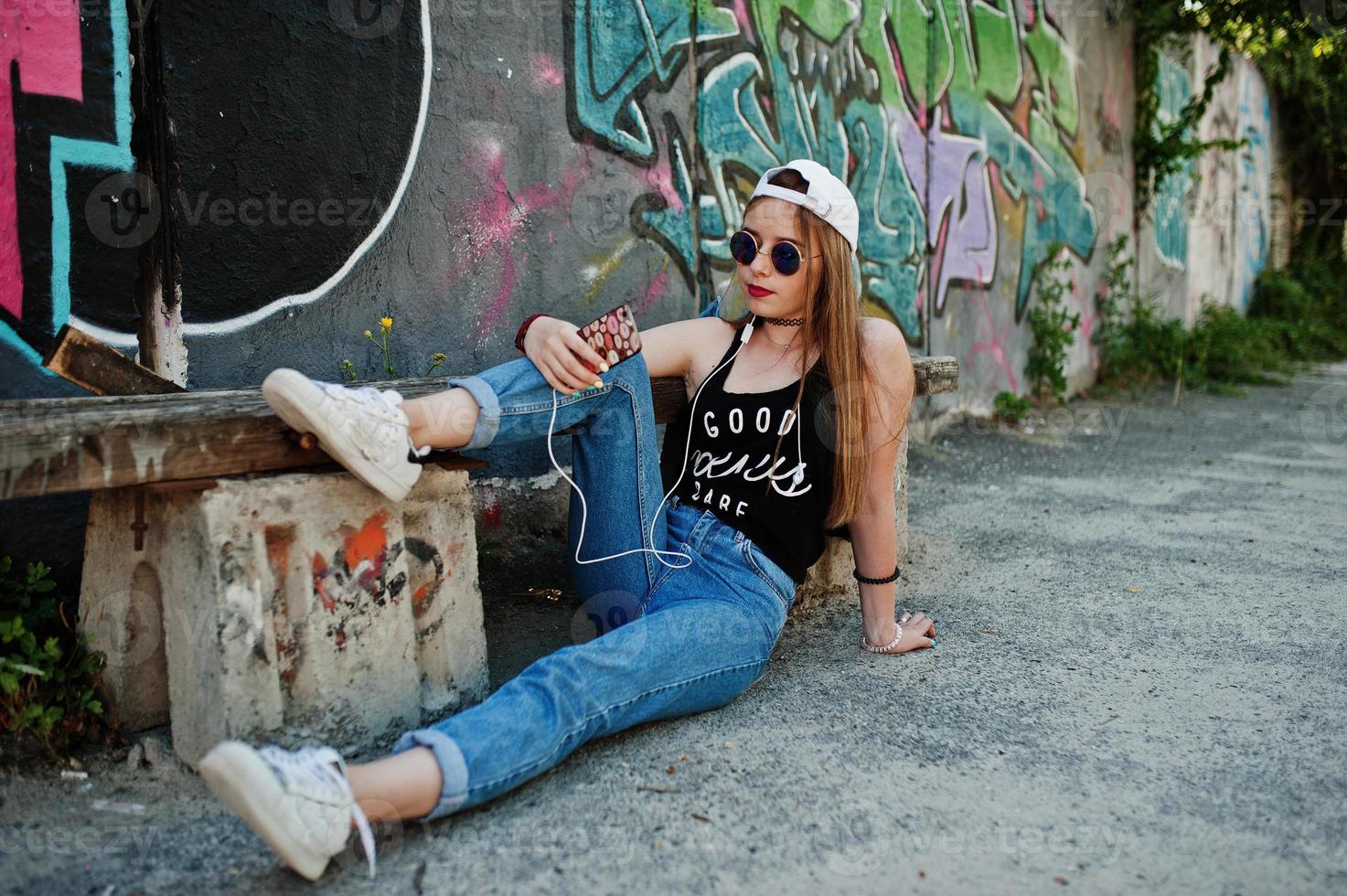 menina elegante casual hipster no boné, óculos escuros e calça jeans, ouvindo música de fones de ouvido do telefone celular contra grande parede de graffiti. foto