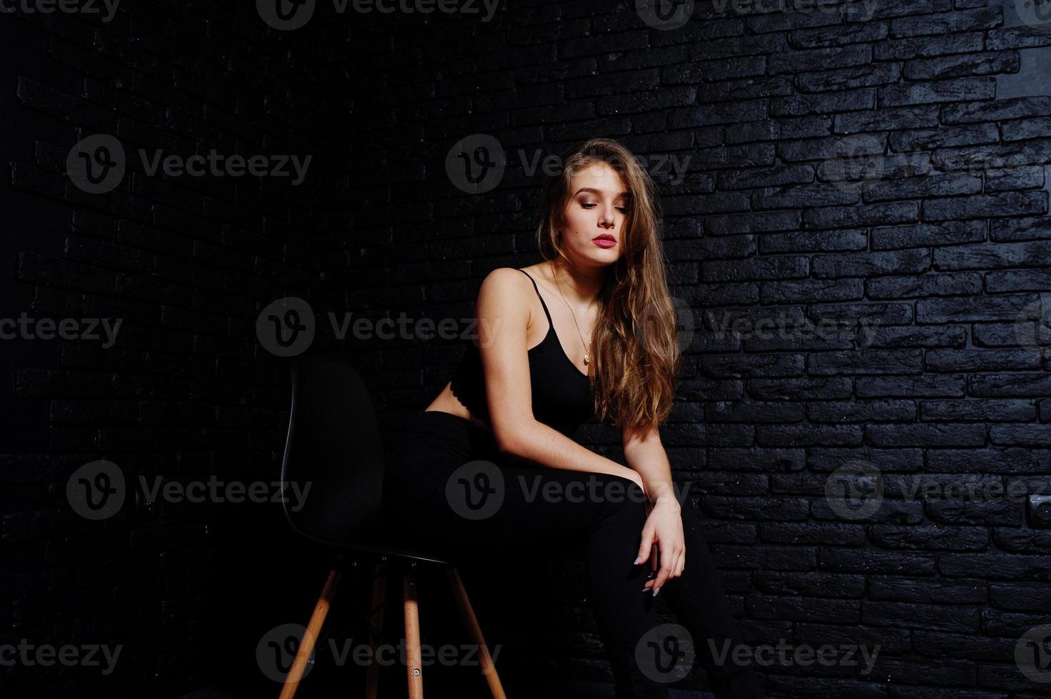 menina morena bonita usa preto, sentado e posando na cadeira no estúdio contra a parede de tijolos escuros. retrato de modelo de estúdio. foto