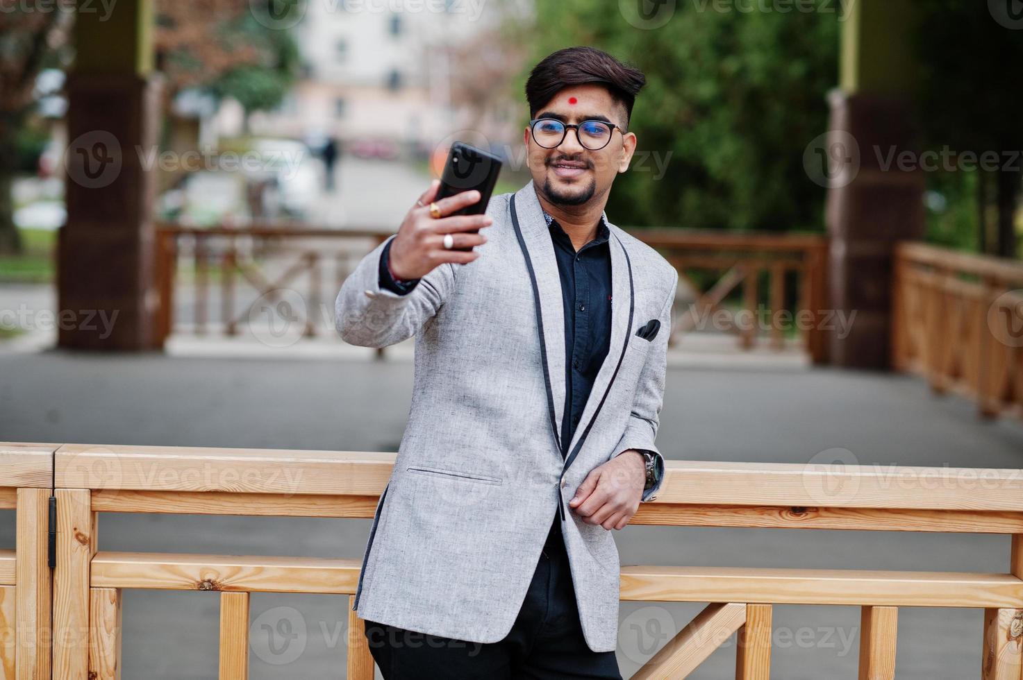 homem de negócios indiano elegante com bindi na testa e óculos, use terno cinza posou ao ar livre e fazendo selfie no celular. foto