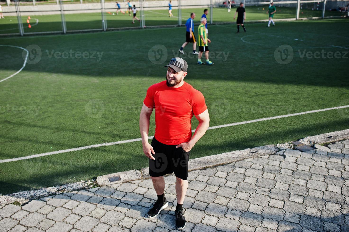 jovem homem musculoso barbudo brutal usar camisa vermelha, shorts e boné no estádio. foto