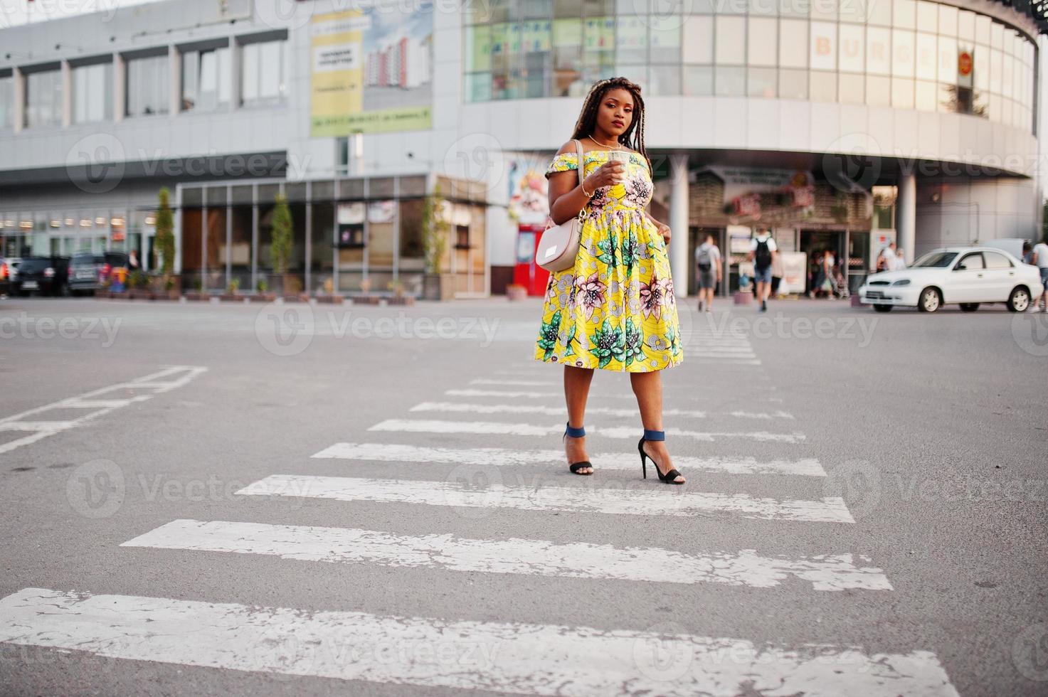 linda garota afro-americana de pequena altura com dreadlocks, use um vestido amarelo colorido, andando na faixa de pedestres contra o centro comercial com uma xícara de café à mão. foto