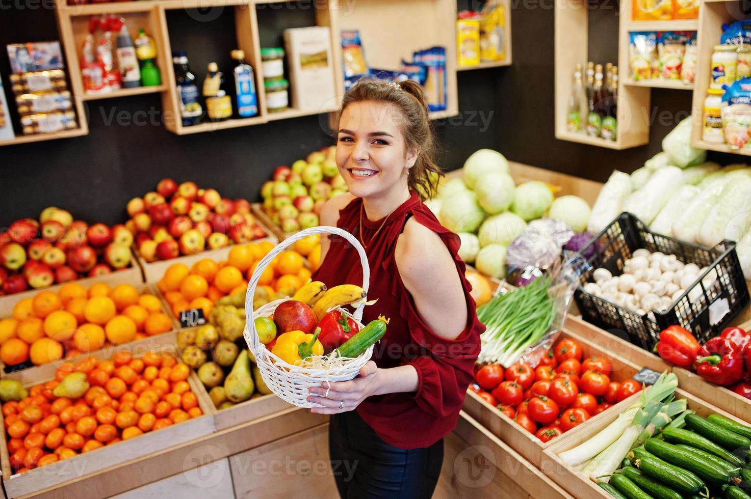 garota de vermelho segurando diferentes frutas e legumes na cesta na loja de frutas. foto