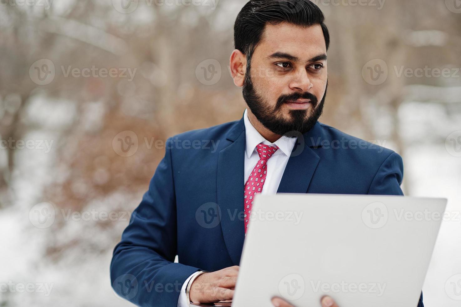 homem de negócios de barba indiana elegante de terno posou em dia de inverno ao ar livre com o laptop nas mãos. foto