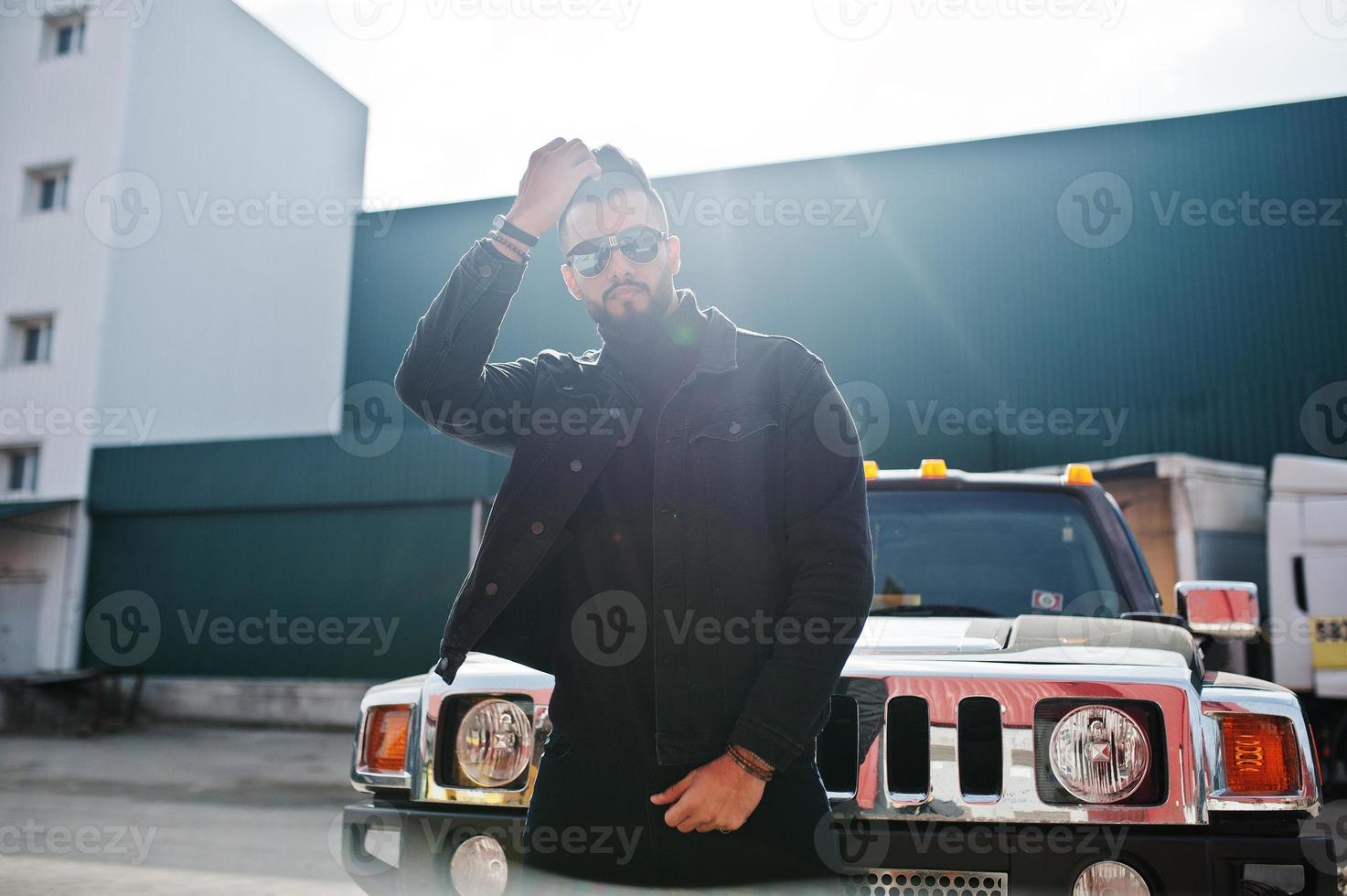 moda barba rica homem árabe usar jaqueta jeans preta e óculos escuros posou contra grande carro suv preto. cara modelo árabe elegante, bem sucedido e elegante. foto