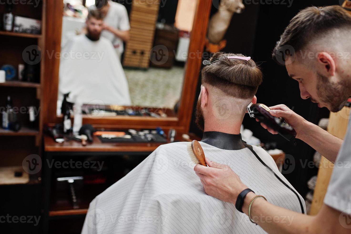 jovem barbudo cortando o cabelo pelo cabeleireiro enquanto está sentado na cadeira na barbearia. alma de barbeiro. foto