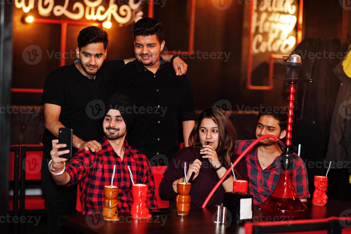 grupo de amigos indianos se divertindo e descansando na boate, bebendo coquetéis e fumando narguilé, olhando para telefones celulares e fazendo selfie. foto