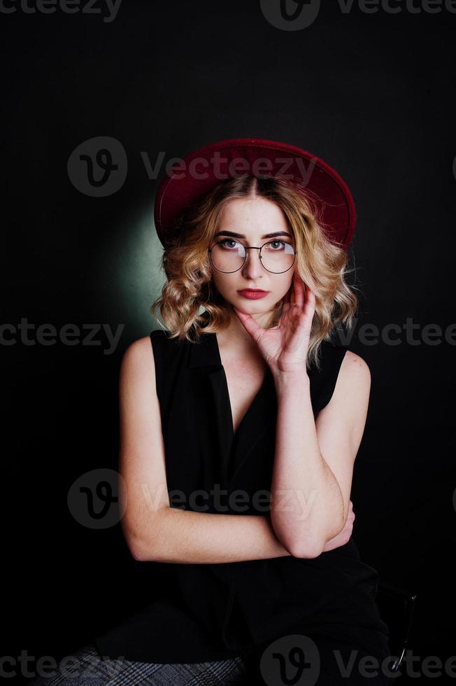 retrato de estúdio de menina loira com roupa preta, chapéu vermelho e óculos contra um fundo escuro. foto