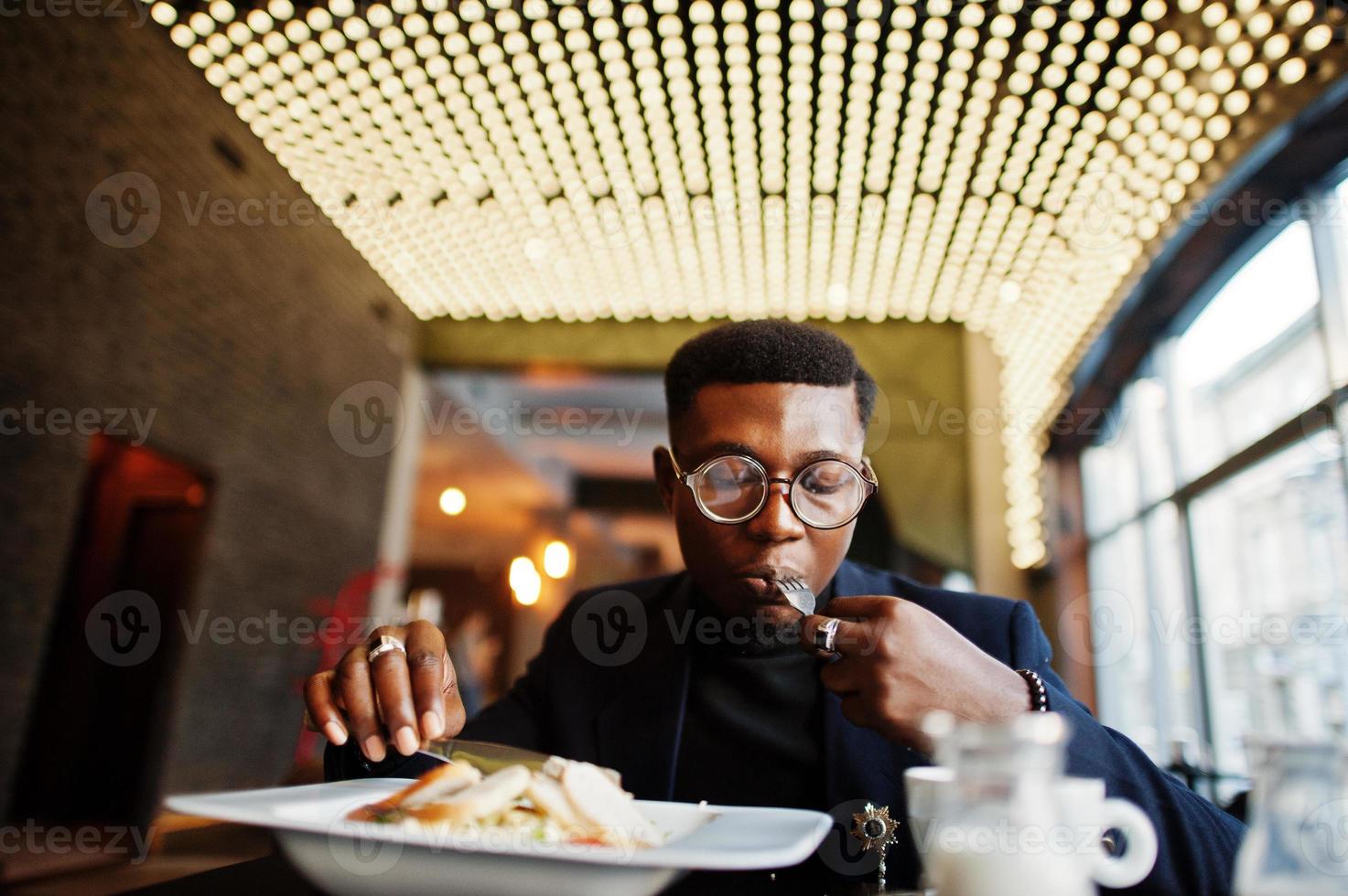 homem afro-americano elegante de terno e óculos sentado no café e comendo salada. foto