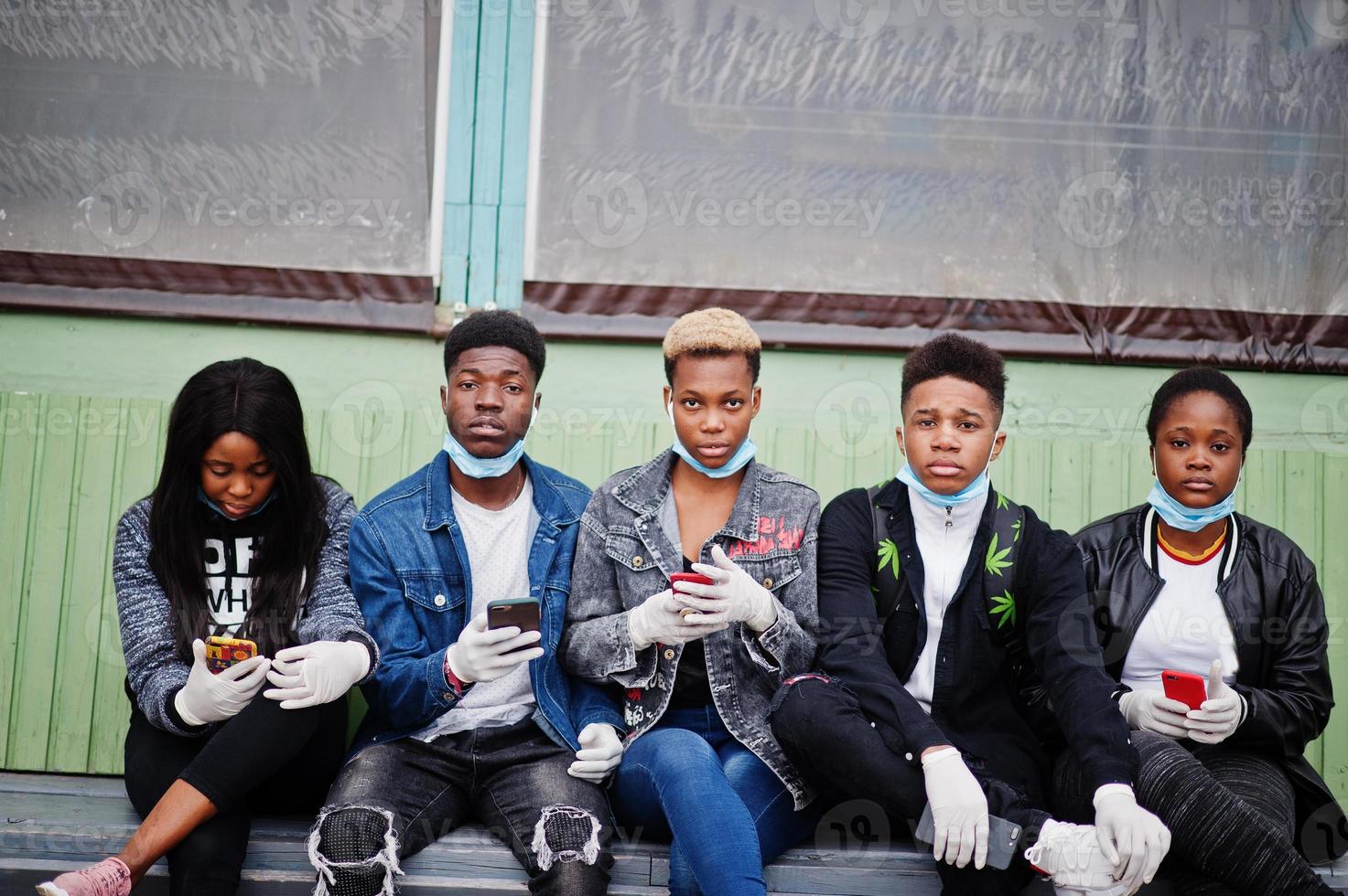 grupo de amigos adolescentes africanos sentados com telefones, usando máscaras médicas protegem contra infecções e doenças quarentena do vírus coronavírus. foto