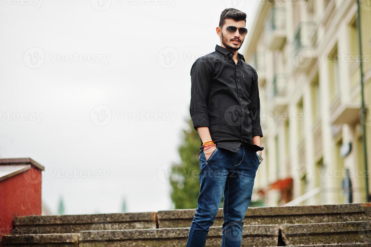 homem elegante indiano na camisa preta e óculos de sol posou ao ar livre. foto