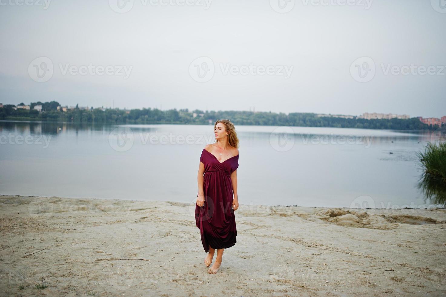 mulher loira sensual descalça no vestido vermelho marsala posando contra o lago na areia. foto