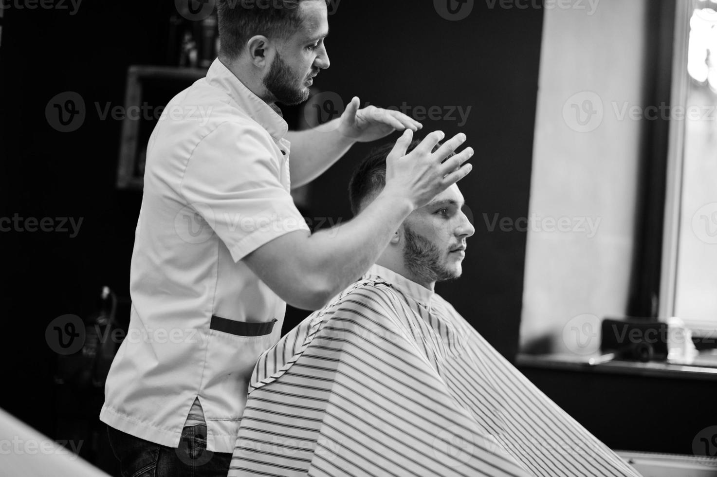 jovem barbudo cortando o cabelo pelo cabeleireiro enquanto está sentado na cadeira na barbearia. alma de barbeiro. foto