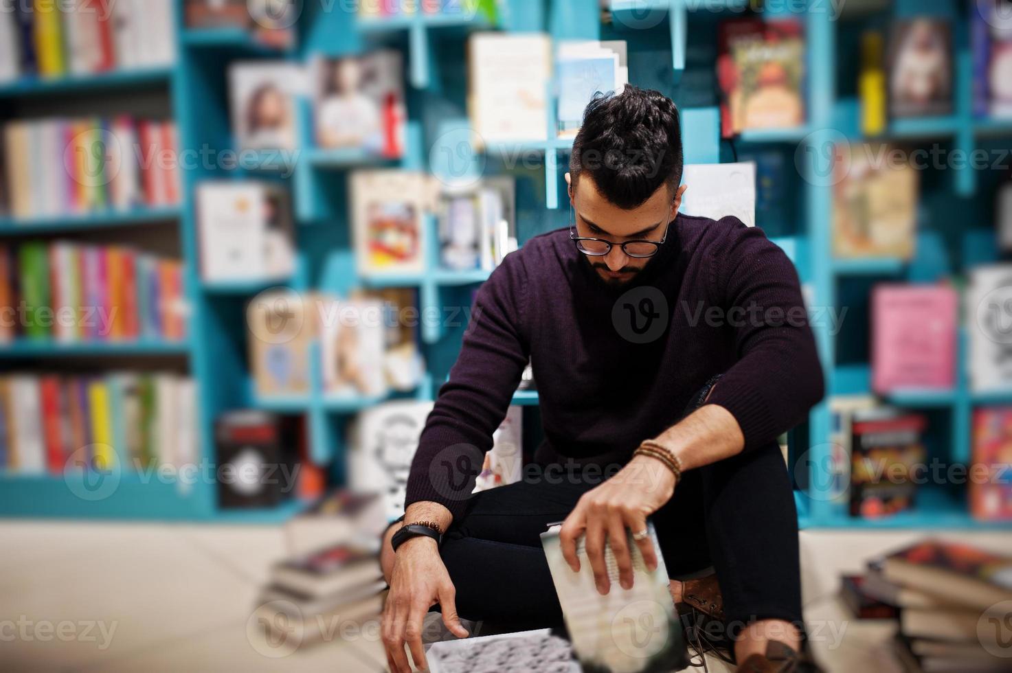 homem alto e inteligente estudante árabe, use gola rulê violeta e óculos, na biblioteca, sentado contra as prateleiras de livros. foto