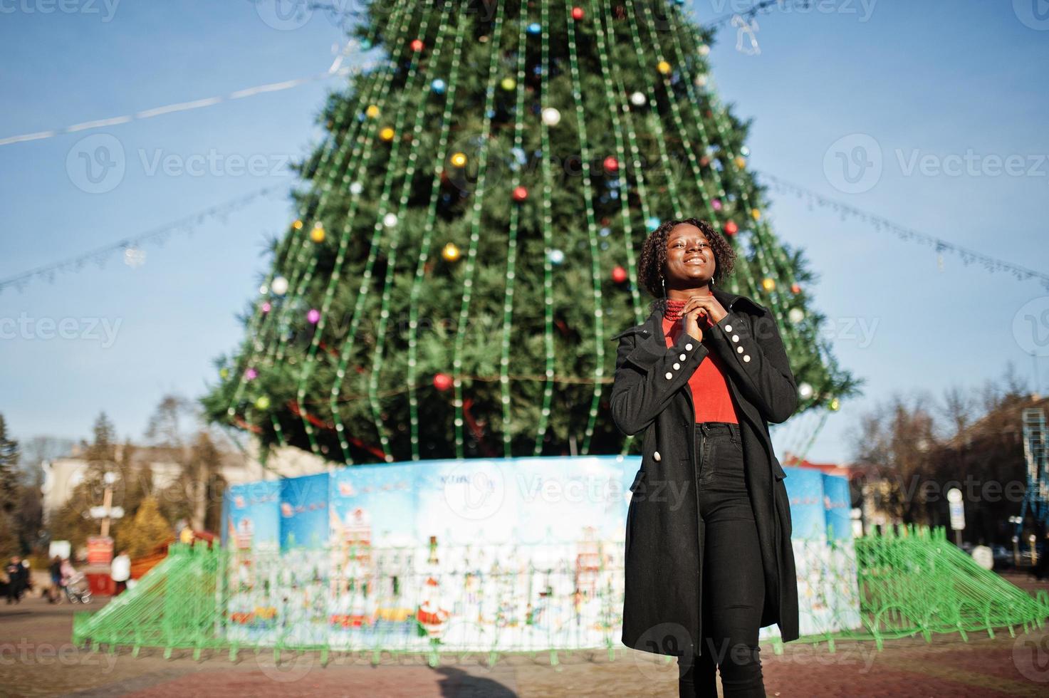 retrato de uma mulher africana de cabelos cacheados vestindo elegante casaco preto e gola alta vermelha posando ao ar livre contra a árvore principal do ano novo da cidade. foto