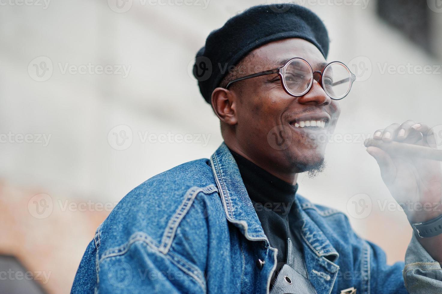 feche o retrato do homem afro-americano na jaqueta jeans, boina e óculos, fumando charuto. foto