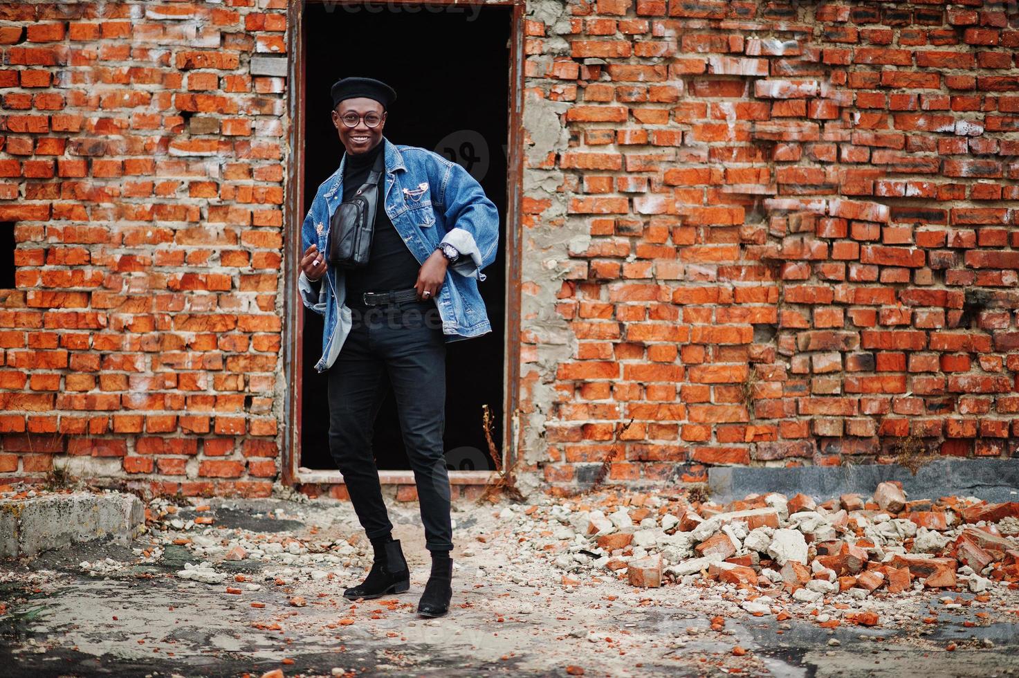 homem afro-americano na jaqueta jeans, boina e óculos contra a parede de tijolos no telhado abandonado. foto