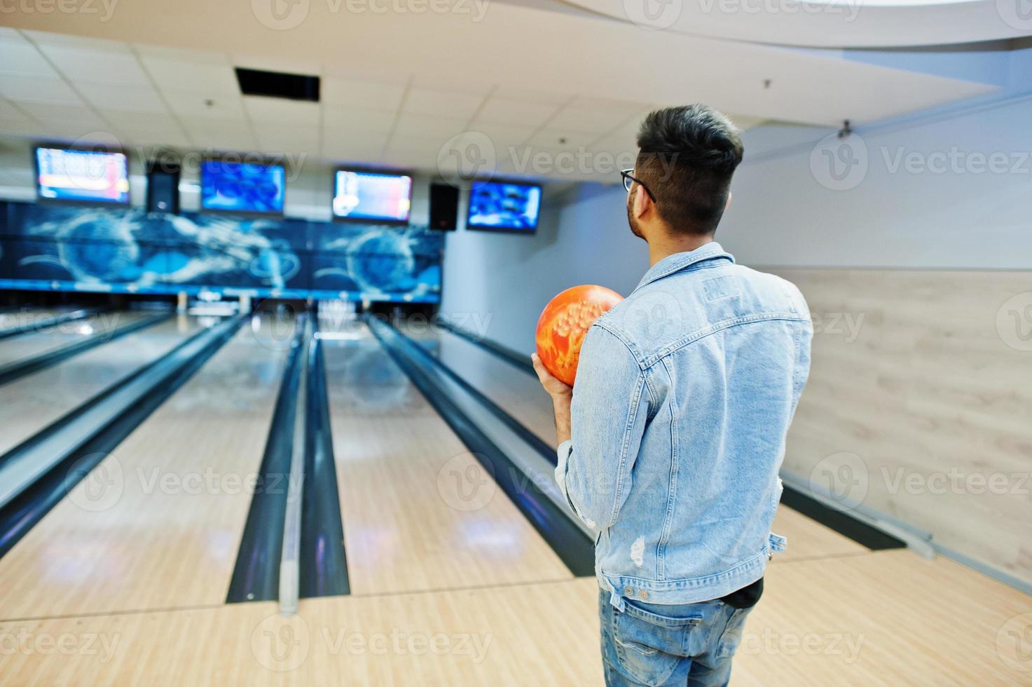 parte traseira do homem asiático elegante na jaqueta jeans e óculos em pé na pista de boliche com bola na mão. foto