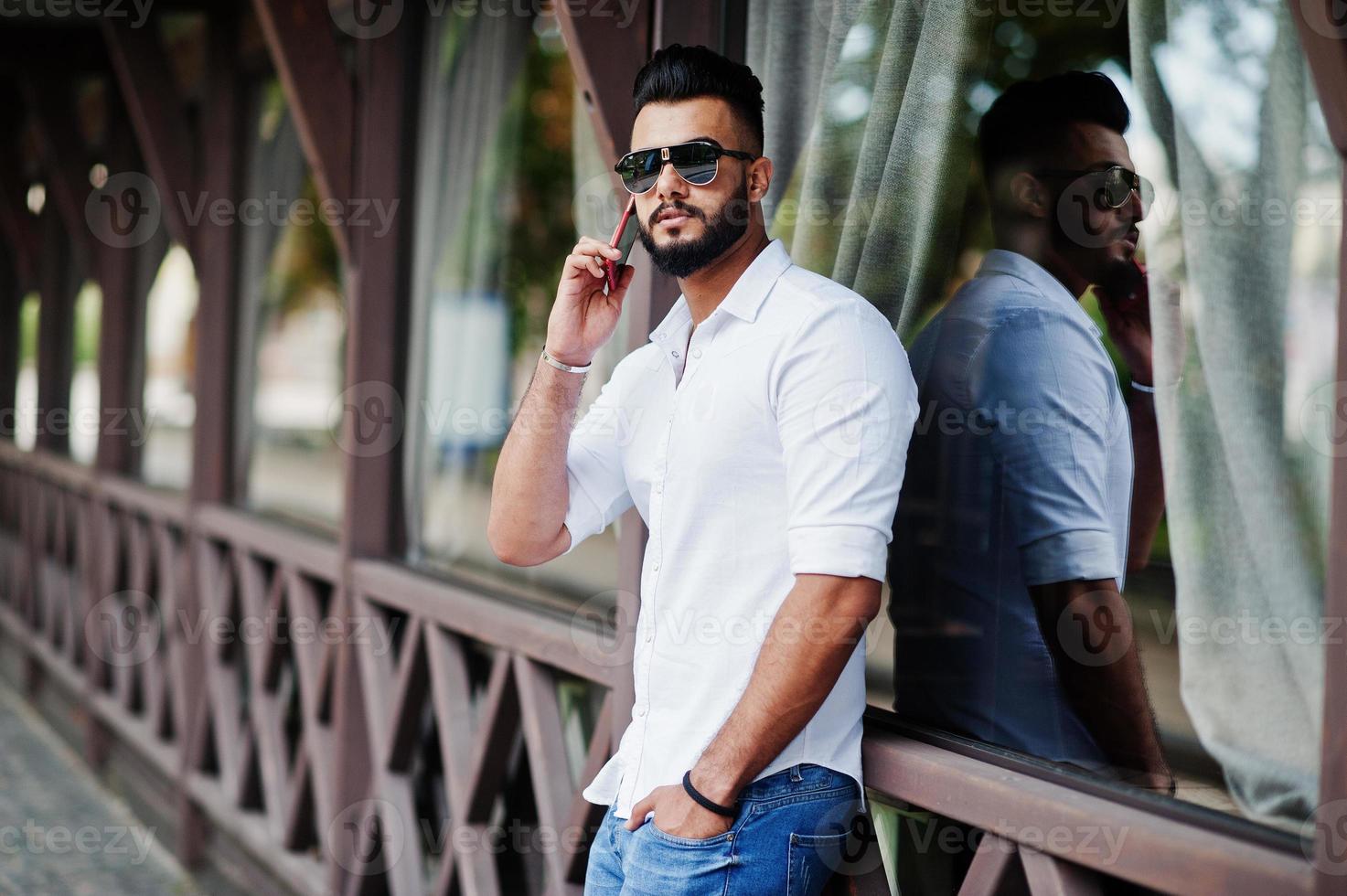 elegante modelo de homem árabe alto na camisa branca, jeans e óculos de sol posou na rua da cidade. barba cara árabe atraente falando no celular. foto